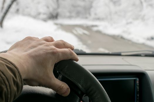 Man's hand on the steering wheel of a car that moves in the snowy forest on a wet slushy road