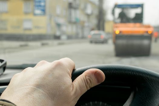 Close-up, the driver's hand on the steering wheel of the car on the background of construction road machinery, road paver and road repair in cloudy weather