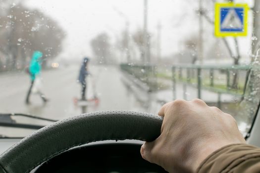 Close-up, the driver's hand on the steering wheel of the car on the background of a pedestrian crossing in rainy weather and a mother with a child on a scooter
