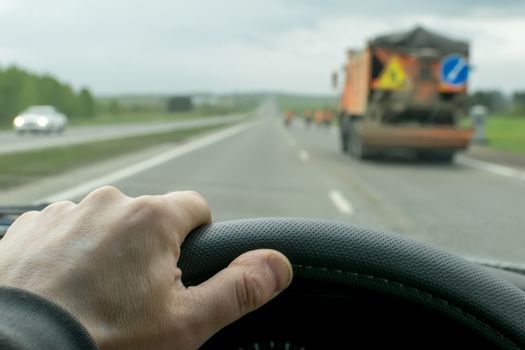 The hand of the driver who moves on a country road in cloudy weather in the evening on the car against the background of special equipment for repair of the road with warning signs