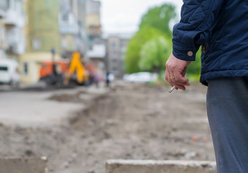 Hand man with a cigarette on the background of the destroyed and repair the road in the yard of a house with construction machinery and machines