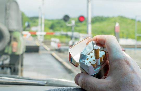 View of the driver hand with a pack of cigarettes on the steering wheel of the car, which stopped before a closed railway crossing at a red light