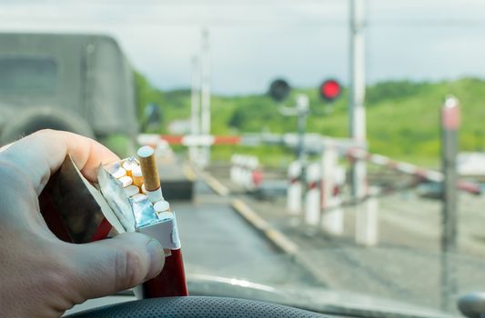 View of the driver hand with a pack of cigarettes on the steering wheel of the car, which stopped before a closed railway crossing at a red light