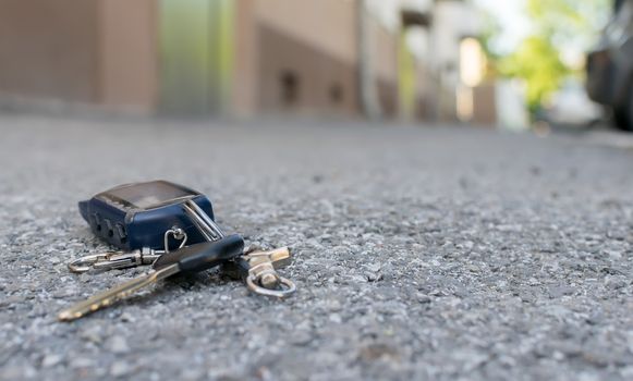 The lost keychain, car alarm remote, lies on the asphalted sidewalk of the road, near an entrance of a house with the parked cars