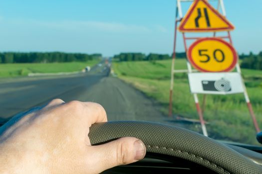 The driver's hand on the steering wheel of the car on the background of a suburban highway on the background of a road sign with speed limit and a warning about the camera photo and video recording