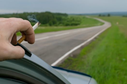 the sight of a people hand leaning on a car, holding a Smoking cigarette against a hilly landscape and the road going away