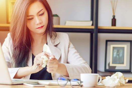 Asian businesswoman stressed with unsuccessful work, squeeze paper in hand sitting at office desk covered with crumpled paper and feeling tired after work fail, give up, no idea