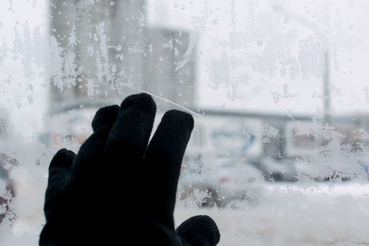 the hand of a man in a winter glove touches the icy glass of the car, on which snow patterns, snowflakes, on the background of the cityscape. Cold in the car