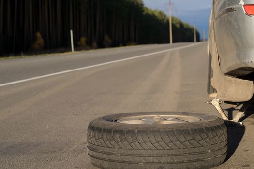 Spare wheel lying near the car. The car is mounted on the Jack. View asphalt road at sunset