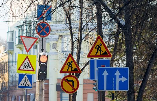 a large number of road signs, symbols of detours, road repairs near the traffic light with a pedestrian crossing on the background of city buildings and tree branches