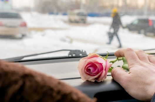 the hand of the driver of the car reaches for a scarlet rose flower lying on the dashboard of the car, against the background of a girl walking along a winter snowy street