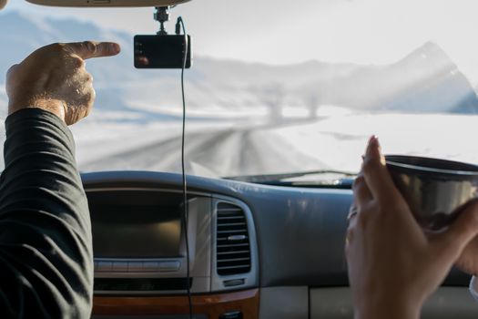 The hand of a man, the driver, shows the girl with a mug of tea, the direction of the road, in winter, in the snow-covered mountain area