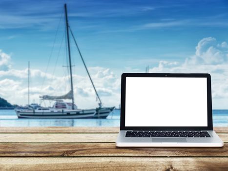 computer laptop with blank white screen on wooden table with yacht boats floating on the sea at background. Travel vacation concept.