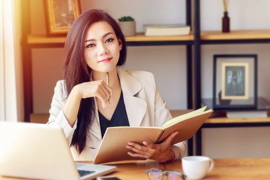 portrait of beautiful and confident Asian business woman in working with notebook laptop and book manage job work at workplace, look at camera and smile. happy businesswoman concept