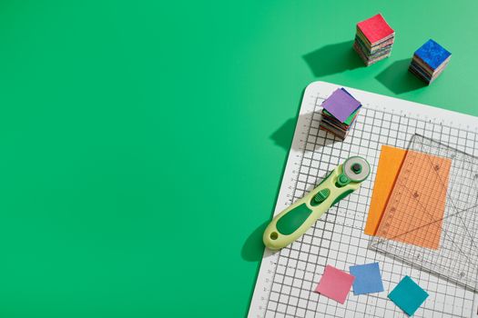 Rotary cutter, ruler, bright square pieces of fabric, stacks of bright fabric square pieces on craft mat, green background