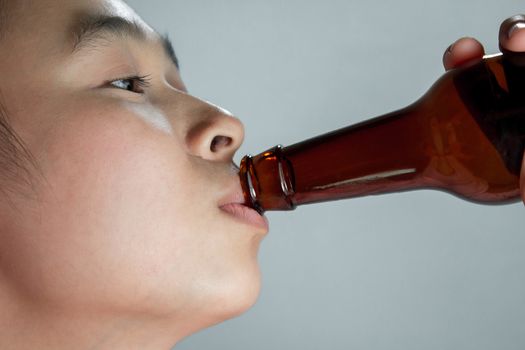 Close-up of Asian young woman drinking beer from bottle on grey background.