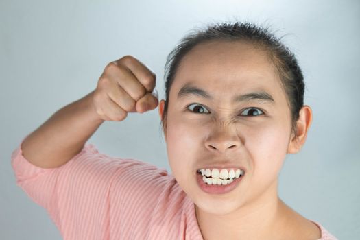 Portrait angry face of Asian young woman in pink shirt and raising fist frustrated on grey background.