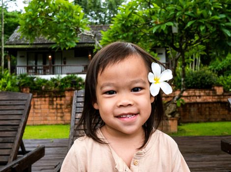 Portrait of Asian little child girl with white plumeria flower on the ear and smile face sitting on wooden chair  nearing swimming pool.