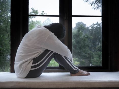 Portrait of sad asian woman sitting hugging the knee on window sill in a rainy day at home.