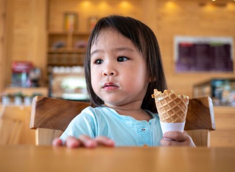 Portrait of Asian little child girl eating ice cream in cone deliciously in cafe.