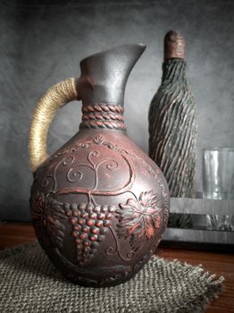 Ceramic wine jug made of red clay with a braided handle, handmade. There is a wine bottle and a glass next to it.