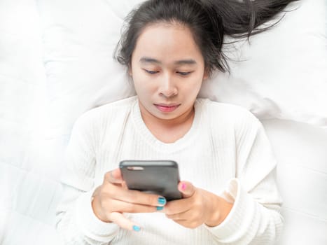 Asian woman reading good news on mobile phone during rest on bed at home. Technology and relaxation concept.