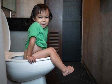 Adorable Asian child girl sitting on toilet bowl at home in the morning. Health care concept.