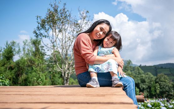 Happy mother sitting with her daughter on wooden ground over trees  with mountains background under blue sky in summer day.