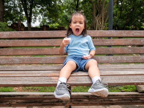 Happy Asian little child girl sitting on wooden chair in the park.