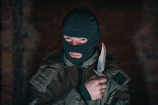 Suspicious look of a bandit in a balaclava with a knife on a background of a brick wall in the dark