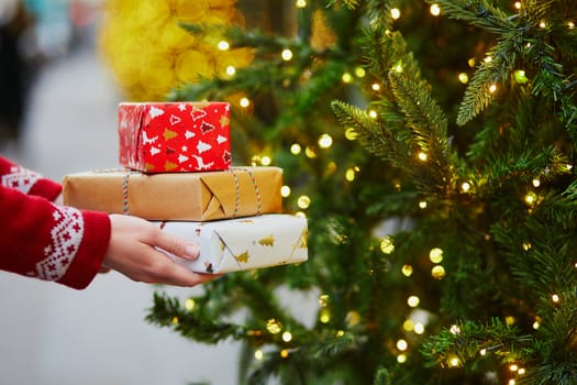 Woman hands holding pile of Christmas presents near New year tree decorated with lights and beads