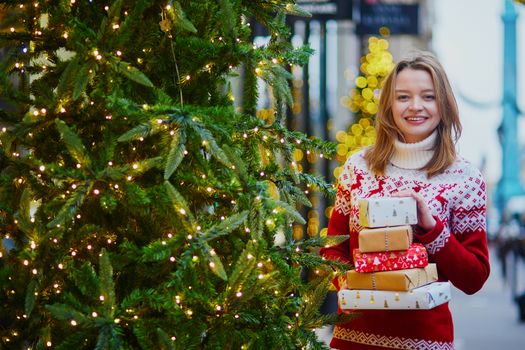 Happy young girl in warm red knitted sweater with pile of holiday gifts on a street of Paris decorated for Christmas