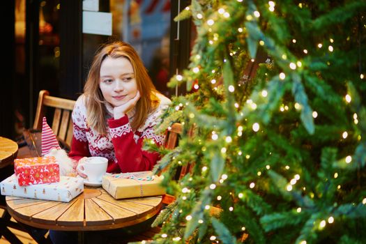 Cheerful young girl in warm red knitted holiday sweater drinking coffee or hot chocolate in cafe decorated for Christmas and unwrapping Christmas present