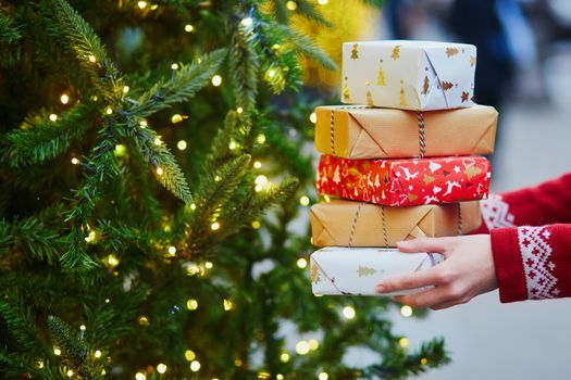 Woman hands holding pile of Christmas presents near New year tree decorated with lights and beads