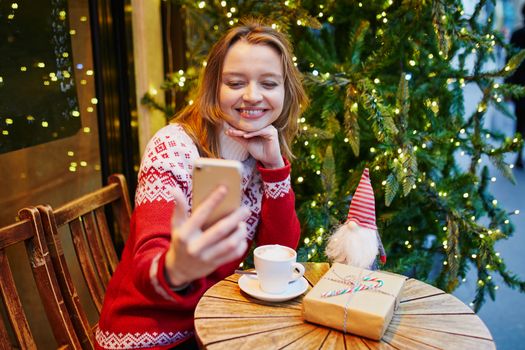 Cheerful young girl in holiday sweater in cafe decorated for Christmas, making selfie with her coffee cup and Christmas present