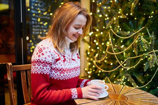 Cheerful young girl in holiday sweater drinking coffee or hot chocolate in cafe decorated for Christmas