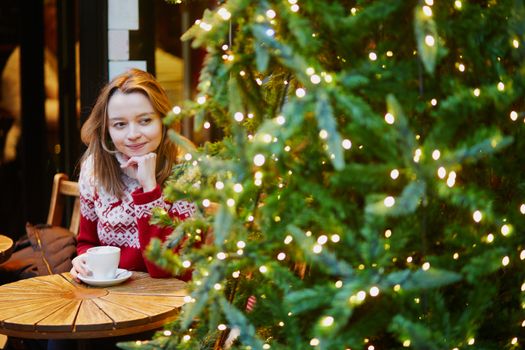 Cheerful young girl in holiday sweater drinking coffee or hot chocolate in cafe decorated for Christmas