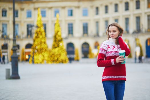 Happy young girl in warm red knitted holiday sweater walking with hot drink to go on a street of Paris decorated for Christmas