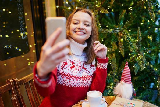 Cheerful young girl in holiday sweater in cafe decorated for Christmas, making selfie with her coffee cup and Christmas present