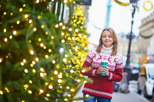 Happy young girl in warm knitted holiday sweater walking with hot drink to go on a street of Paris decorated for Christmas