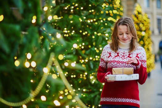 Happy young girl in holiday sweater with pile of holiday gifts on a street of Paris decorated for Christmas