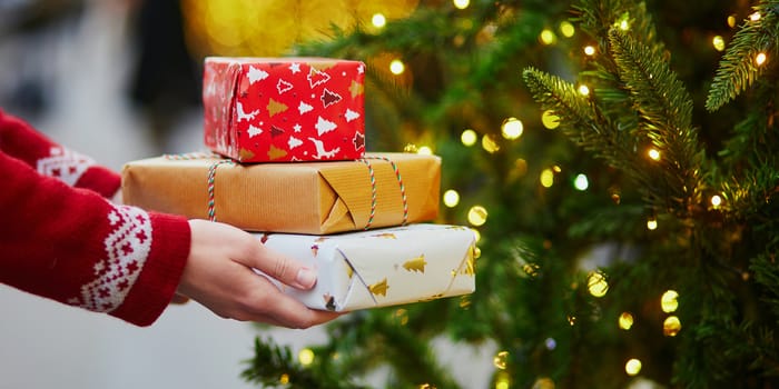 Woman hands holding pile of Christmas presents near New year tree decorated with lights and beads