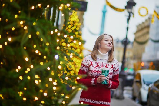 Happy young girl in holiday sweater walking with hot drink to go on a street of Paris decorated for Christmas