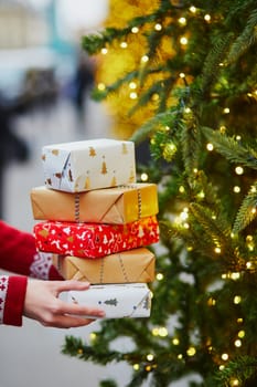 Woman hands holding pile of Christmas presents near New year tree decorated with lights and beads