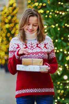 Happy young girl in warm red knitted sweater with pile of holiday gifts on a street of Paris decorated for Christmas