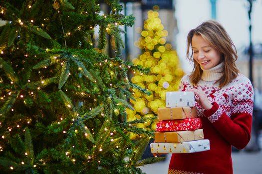 Happy young girl in warm red knitted sweater with pile of holiday gifts on a street of Paris decorated for Christmas