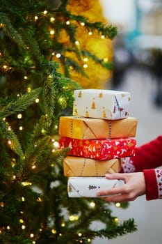 Woman hands holding pile of Christmas presents near New year tree decorated with lights and beads