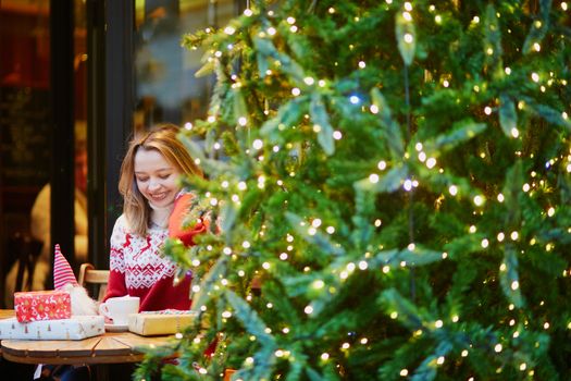 Cheerful young girl in warm red knitted holiday sweater drinking coffee or hot chocolate in cafe decorated for Christmas and unwrapping Christmas present