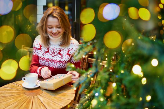 Cheerful young girl in holiday sweater drinking coffee or hot chocolate in cafe decorated for Christmas and unwrapping Christmas present
