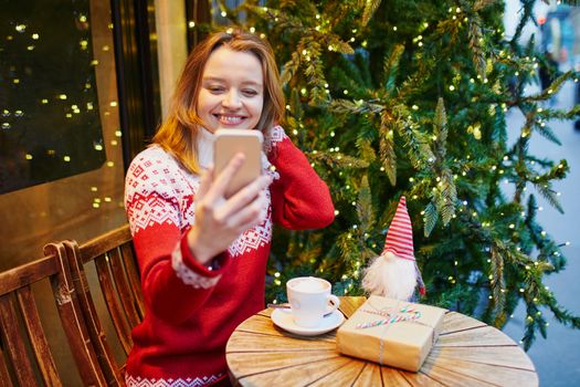 Cheerful young girl in warm red knitted holiday sweater in cafe decorated for Christmas, making selfie with her coffee cup and Christmas present